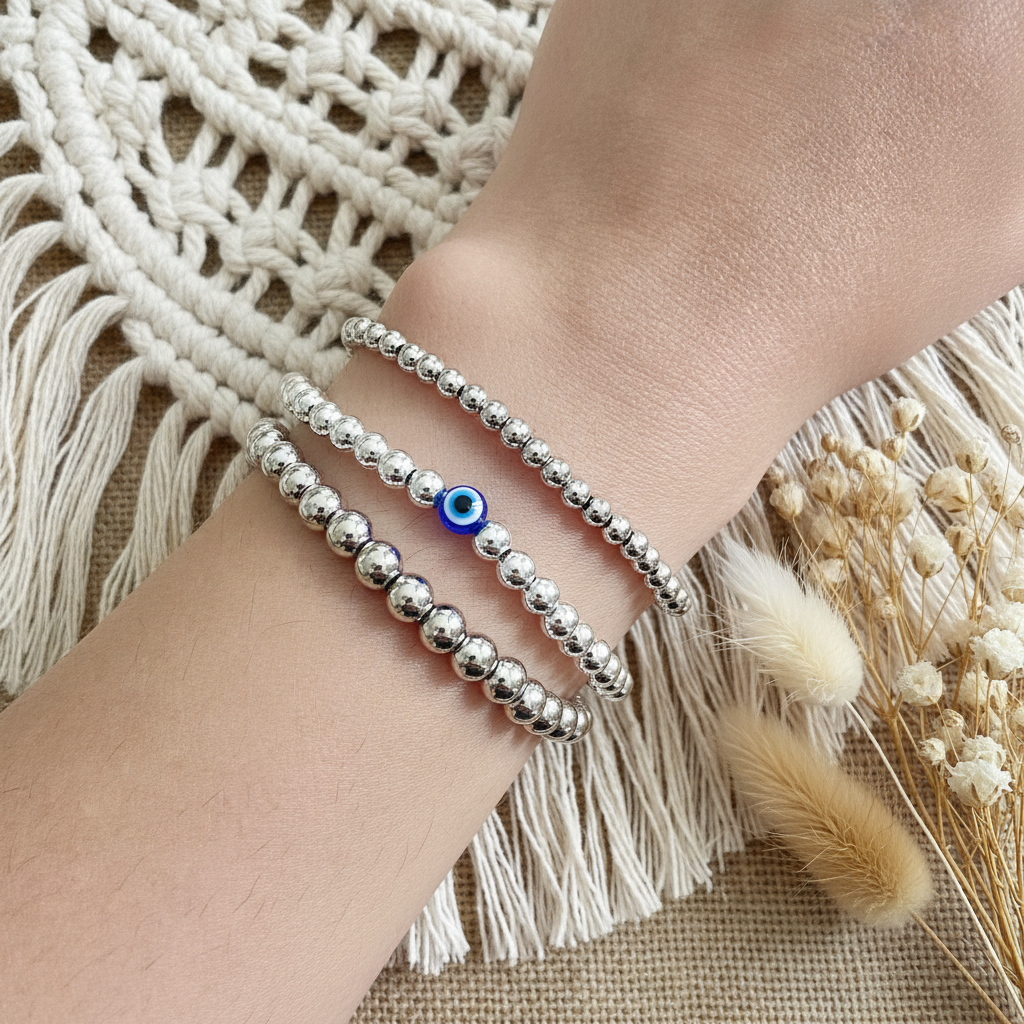 Silver beaded bracelet with a blue evil eye charm on a wrist, against a textured fabric background.