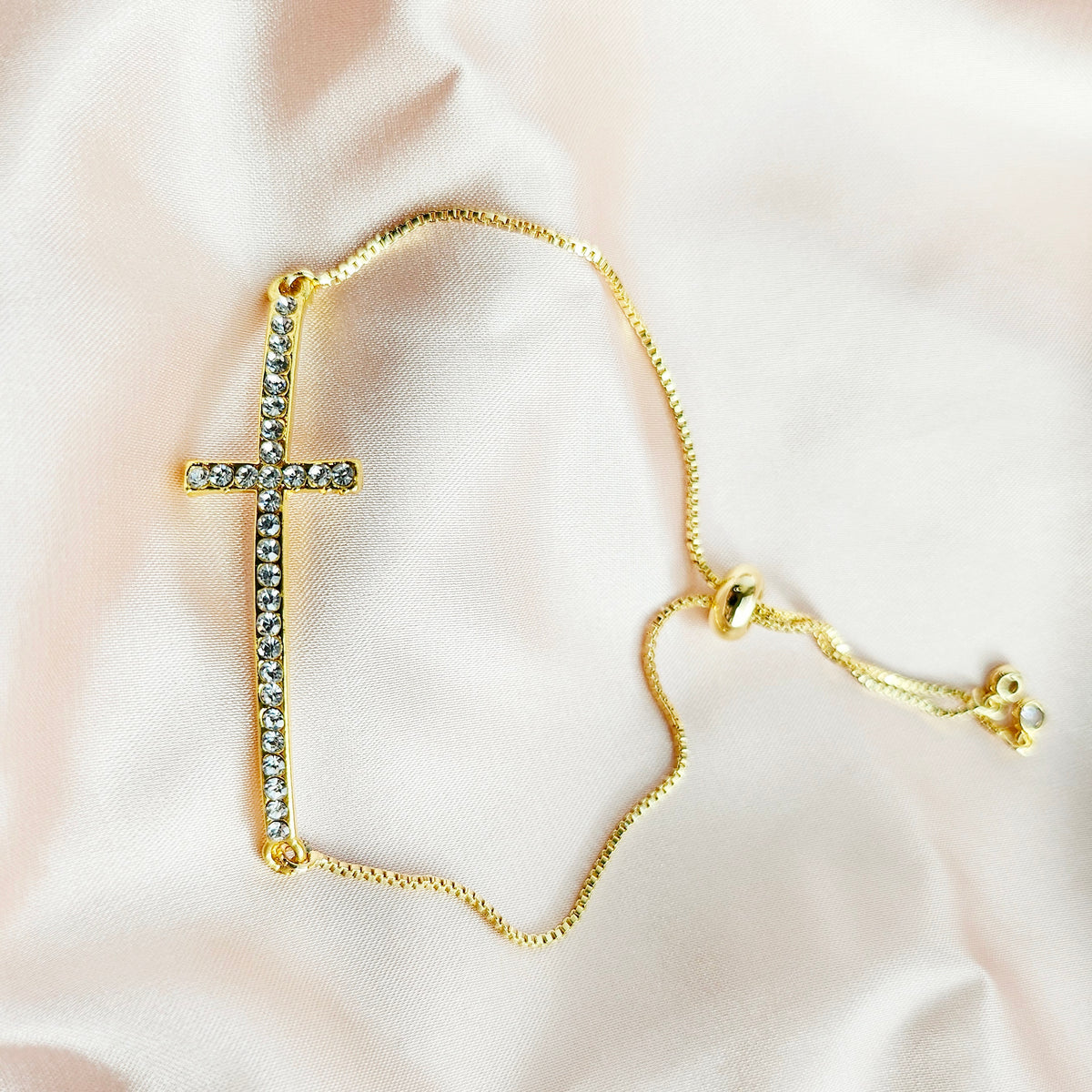 Gold bracelet with a cross pendant on a soft pink fabric background